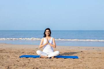 Fototapeta na wymiar Beautiful indian woman wearing white cloths sitting on fitness mat in lotus position meditating at beach. Healthy Life Concept.Copy space.