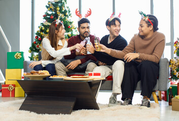 Millennial Asian Indian cheerful happy male female friends holding beer beverage bottles cheers toasting together in living room celebrating Christmas Eve and New Year with full decor Xmas tree