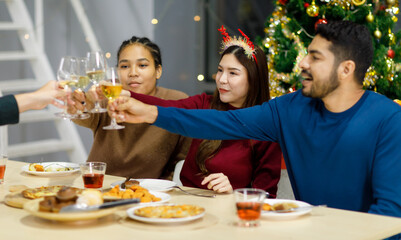 Group of Asian Indian happy cheerful male female friends wears reindeer antlers headband cheers toasting champagne beverage glasses together on dinner table celebrating Christmas eve in dining room