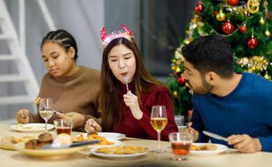 Group of Asian Indian happy cheerful male female friends wears reindeer antlers headband eating food with champagne beverage glasses together on dinner table celebrating Christmas eve in dining room