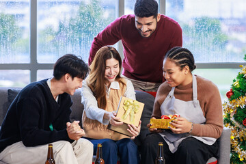 Obraz premium Millennial Asian surprised excited woman model holding opening wrapped present gift box with cheerful male female friends looking cheering in living room celebrating Christmas Eve and New Year
