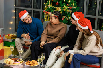 Asian Indian male female friends wear reindeer antlers headband red Santa Claus hat sitting on sofa...