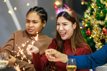 Asian Indian male female friends wears reindeer antlers headband sitting holding playing small sparkler firework celebrating together at dinner table full of food snack in dining room with Xmas tree