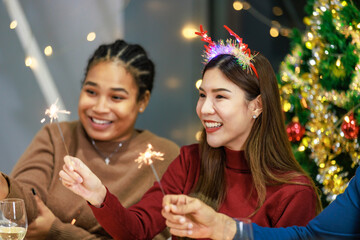 Obraz na płótnie Canvas Asian Indian male female friends wears reindeer antlers headband sitting holding playing small sparkler firework celebrating together at dinner table full of food snack in dining room with Xmas tree