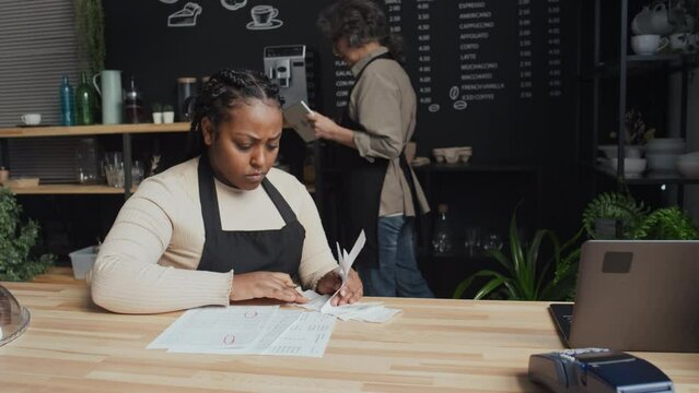 Medium shot of young woman with braids sitting behind counter reconciling accounts while her older colleague checking baristas equipment in background