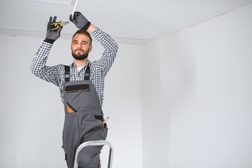 Repairman or professional electrician in workwear installing light spots, standing on the ladder in the white living room - obrazy, fototapety, plakaty