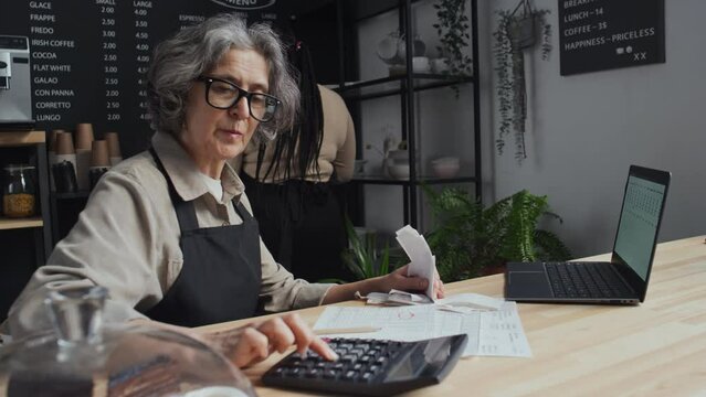 Medium shot of elderly coffee shop worker with glasses reconciling accounts while sitting at counter