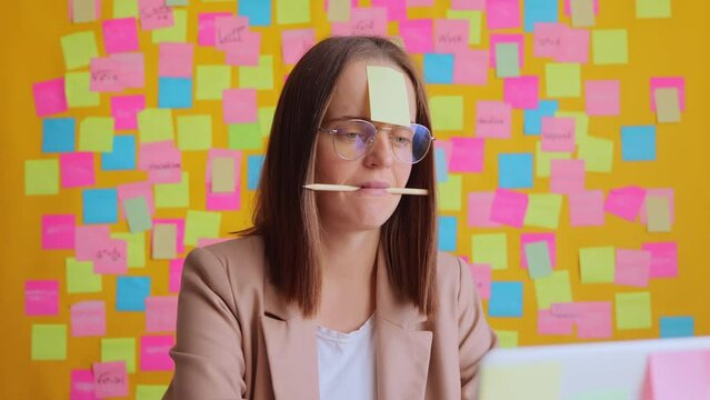Funny woman working on computer against yellow wall with colorful stickers sitting at her workplace with sad expression with memo card on forehead and pencil in mouth.