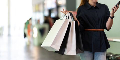Woman using smartphone holding Black Friday shopping bag while standing on the side with the mall