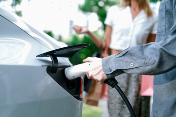 Young couple travel with EV electric car charging in green sustainable city outdoor garden in...