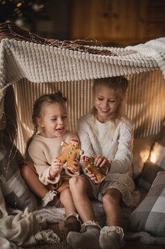 two little girls, blonde sisters in light elegant sweaters and dresses, play at home near the New Year tree in a hut made of blankets. playing hide and seek in a makeshift tent and eating Christmas gi