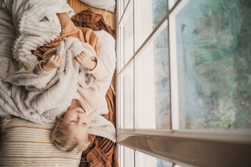 a little girl dozed off on a large wooden windowsill in a warm cozy blanket near the window. children's sleep and disruption of the daily routine. Cozy tender baby. Noise blurred image