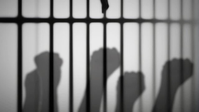 Symbols of a protest in jail: fists raised and moving, silhouetted shapes over the wall of a prison cell, behind bars. Black and white shot.
