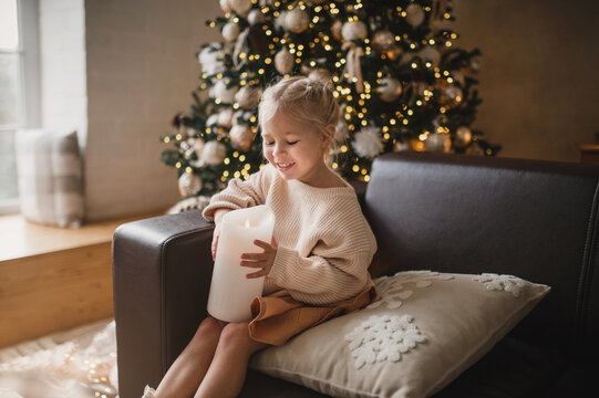A little girl in a light woolen dress poses against the background of a Christmas tree and other New Year decorations with a candle. Noise toned blurred image