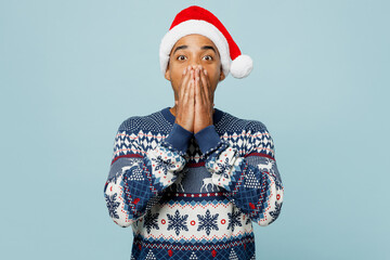 Young surprised shocked astonished man wear knitted sweater Santa hat posing cover mouth with hands...