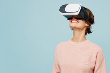 Young smiling happy cheerful woman she wear beige knitted sweater casual clothes watching in vr headset pc gadget isolated on plain pastel light blue cyan background studio portrait Lifestyle concept