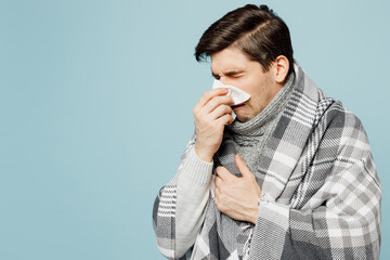 Young sad ill sick man wrapped in gray plaid sneezing use paper napkin for runny nose isolated on...