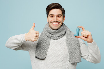 Young ill sick man wear gray sweater scarf use asthma inhaler show thumb up isolated on plain blue...