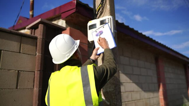 man checks the meter reading electricity in the cottage. An electrical engineer inspects a transformer box and makes entries in a log.