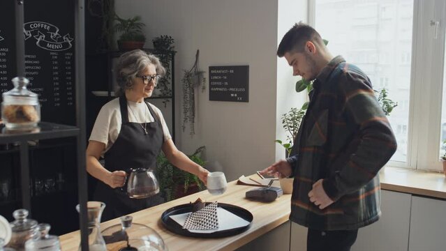 Customer taking his order and paying for it through online app while coffee house worker preparing coffee and baked goods for him