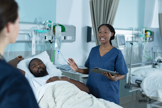 Diverse female doctors and male patient is lying in bed talking in hospital room
