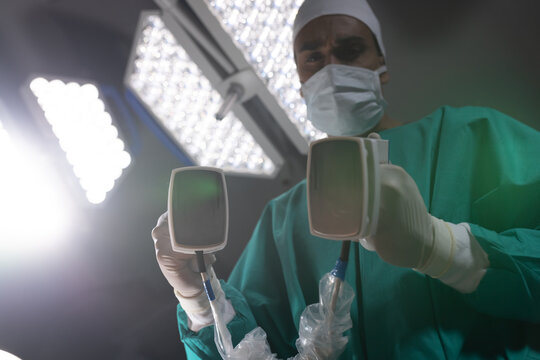 Biracial male surgeon wearing surgical gown using defibrillator in operating theatre at hospital - Powered by Adobe