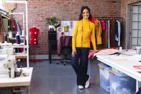 Happy biracial female fashion designer standing with measuring tape and smiling in sunny studio