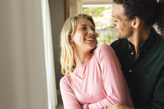 Happy diverse couple embracing and looking at themselves in sunny living room