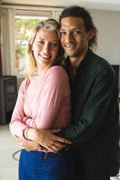 Happy diverse couple embracing, smiling and looking at camera in sunny living room