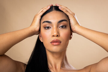 Portrait of biracial woman with dark hair, hands to head and natural make up on brown background