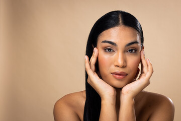 Portrait of biracial woman with dark hair, hands to face and natural make up on brown background