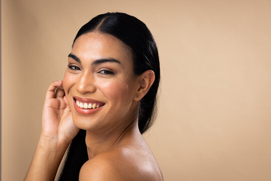Fototapeta Smiling biracial woman with dark hair, hand to face, natural make up on brown background