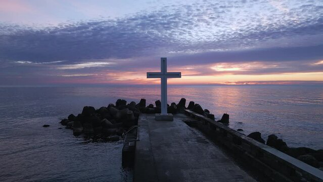 A flight around a Christian Holy cross early in the morning at sunrise. The large cross stands on the edge of a breakwater on the sea coast
