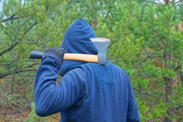 one aggressive man and a bandit in a blue stylish fashionable raglan and a blue hood on his head holds in his hand a small iron new hatchet with a wooden handle behind his back before an attack  
