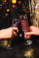  Hands hold glasses of New Year's cocktail with edible glitter and check out among the Christmas lights
