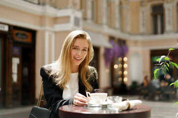 Beautiful  blond woman in stylish   casual business outfit  sitting in cafe   in  beautiful european city.   Enjoing coffee.