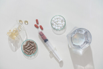 A glass of water, a syringe, medicine displayed on a transparent petri dish and laboratory beaker on a minimalist background. Medicines help support the treatment process.