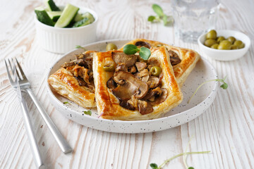 pastry with mushroom and olive filling on puff pastry, white ceramic plate, side bowl with olives, light wooden background, close-up
