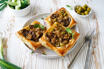 pastry with mushroom and olive filling on puff pastry, white ceramic plate, side bowl with olives, light wooden background, close-up