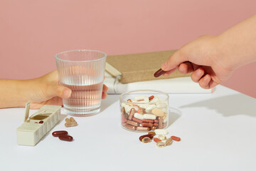 One hand is holding a glass of water, another hand is holding a pill taken from a glass medicine jar. On the table there is a notebook and a weekly pill organizer. Medicine theme.