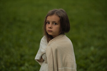 With an enigmatic gaze, a child stands cloaked in a blanket against a green field. This image...