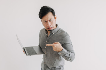 Asian man with beard wear grey shirt, disappointed and unsatisfied face, holding notebook hand pointing at laptop, funny ugly face looking at camera isolated over white background wall.