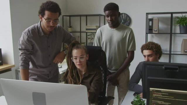 Medium shot of young Caucasian female programmer discussing software on computer with diverse male colleagues while coworking in open space office