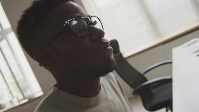 Handheld tilting up shot of young African American male programmer typing on keyboard and looking at computer monitor while developing software in open space minimalist office