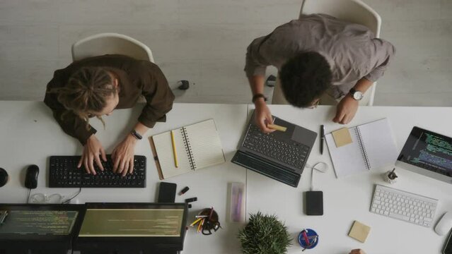 Top view of two professional software developers looking at sticky notes and having discussion while sitting by desktop computers at white office desk working together