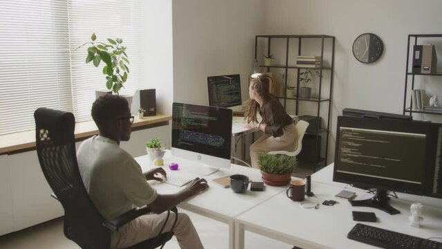 High angle shot of two multiethnic young programmers coding and developing software in open space office. Young Caucasian woman sitting down in chair at work desk in front of desktop computer