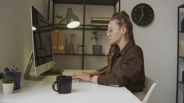 Side view waist up shot of young Caucasian female programmer with dreadlocks writing html code on wireless keyboard sitting in front of computer monitor at workplace in minimalist office
