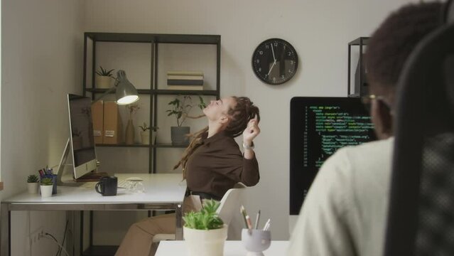 Young tired Caucasian female programmer with dreadlocks stretching her arms and back and doing head rotations at desk while working in open space office with colleague