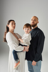 vertical shot of modern stylish parents posing with their son in hands and looking at camera, family
