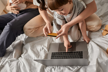 top view of little boy pointing at laptop surrounded by family with his mother holding credit card - obrazy, fototapety, plakaty